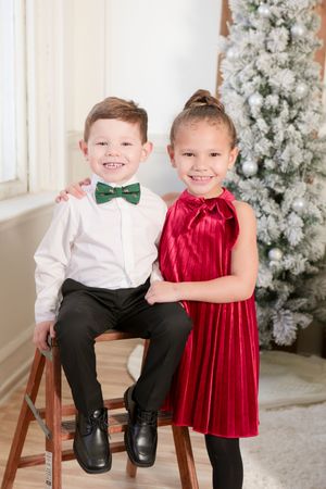Two children in holiday attire pose by a flocked Christmas tree, wearing a green bowtie and red velvet dress.