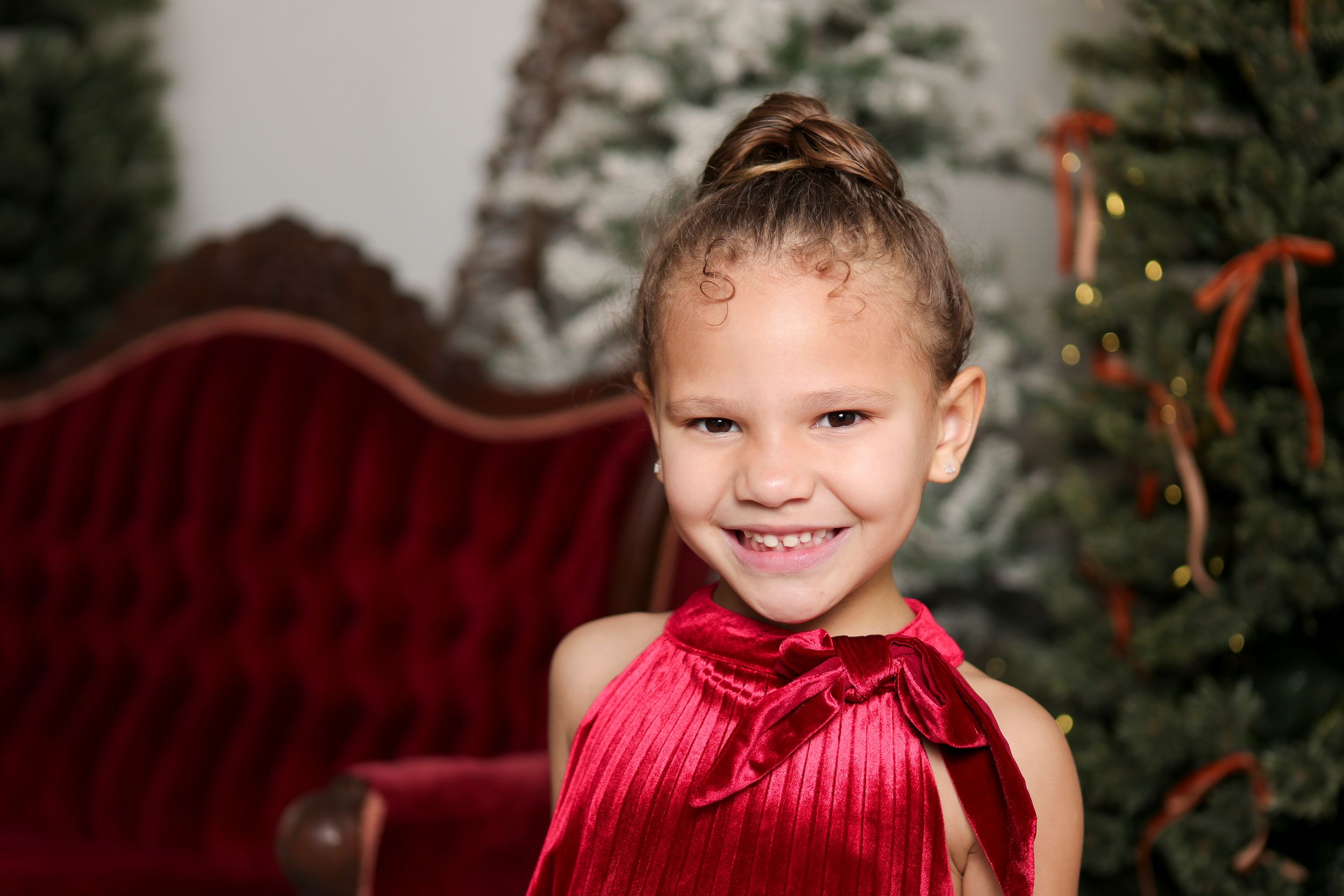 A smiling young person in a bright red holiday dress sits on a velvet couch near a Christmas tree.