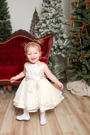 Holiday portrait scene with snowy pine trees, red velvet settee, and child in white party dress on hardwood floor.