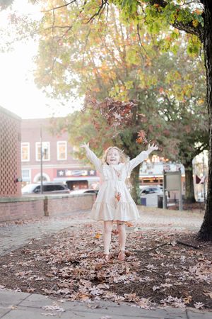 A small child jumping in fallen autumn leaves on a brick sidewalk outside historic brick buildings on a bright fall day.