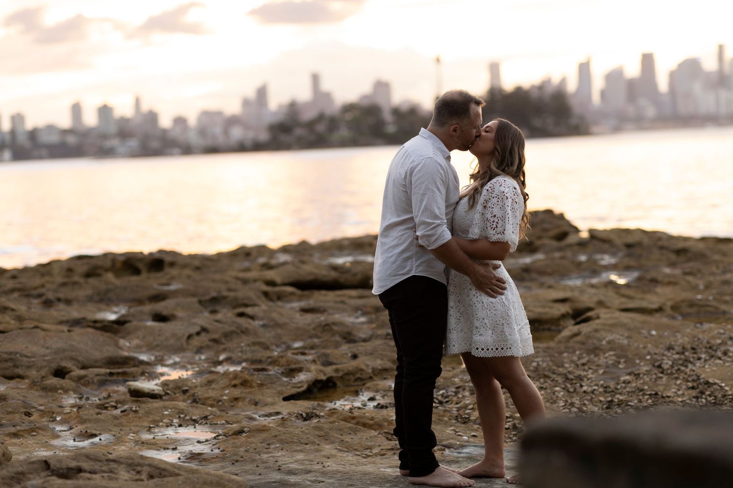 A couple shares an intimate moment on a rocky beach at sunset with a cityscape silhouetted in the background.