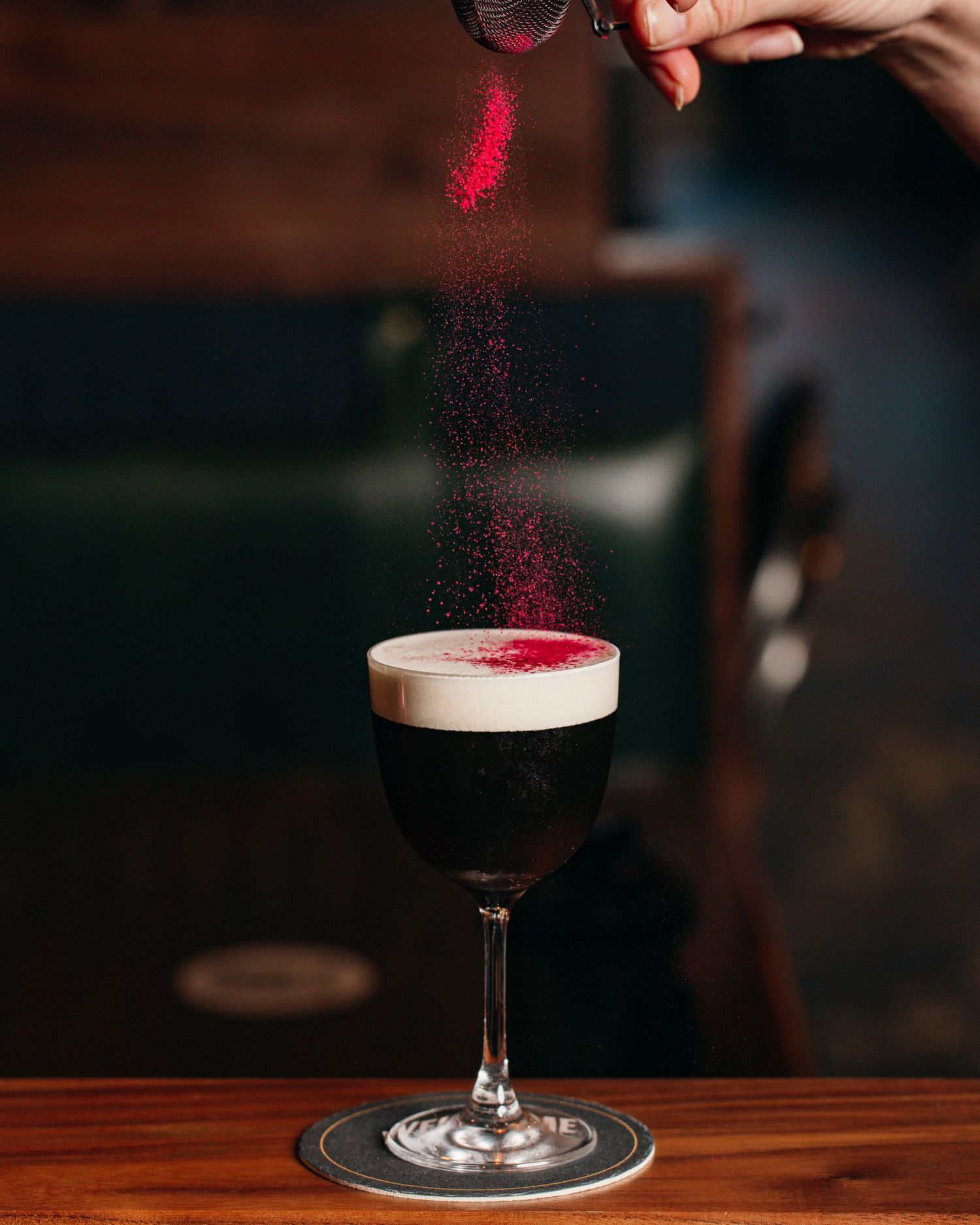 Red powder being sprinkled onto a dark cocktail with frothy top in a stemmed glass.