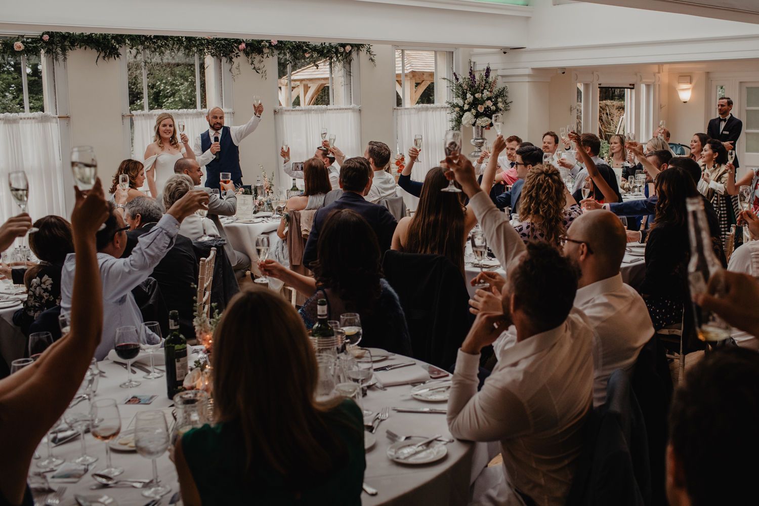 Wedding guests raising glasses in toast during reception speech in bright modern venue with greenery decor.