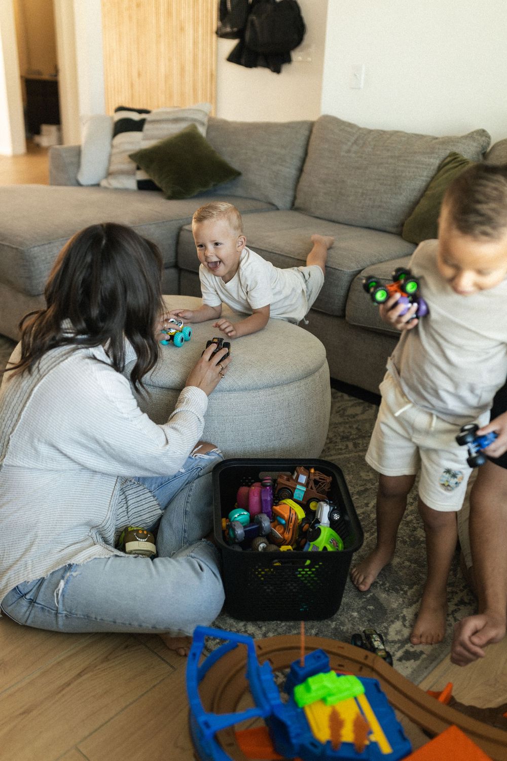 Family playing together with colorful toys scattered on living room floor near grey sectional sofa.