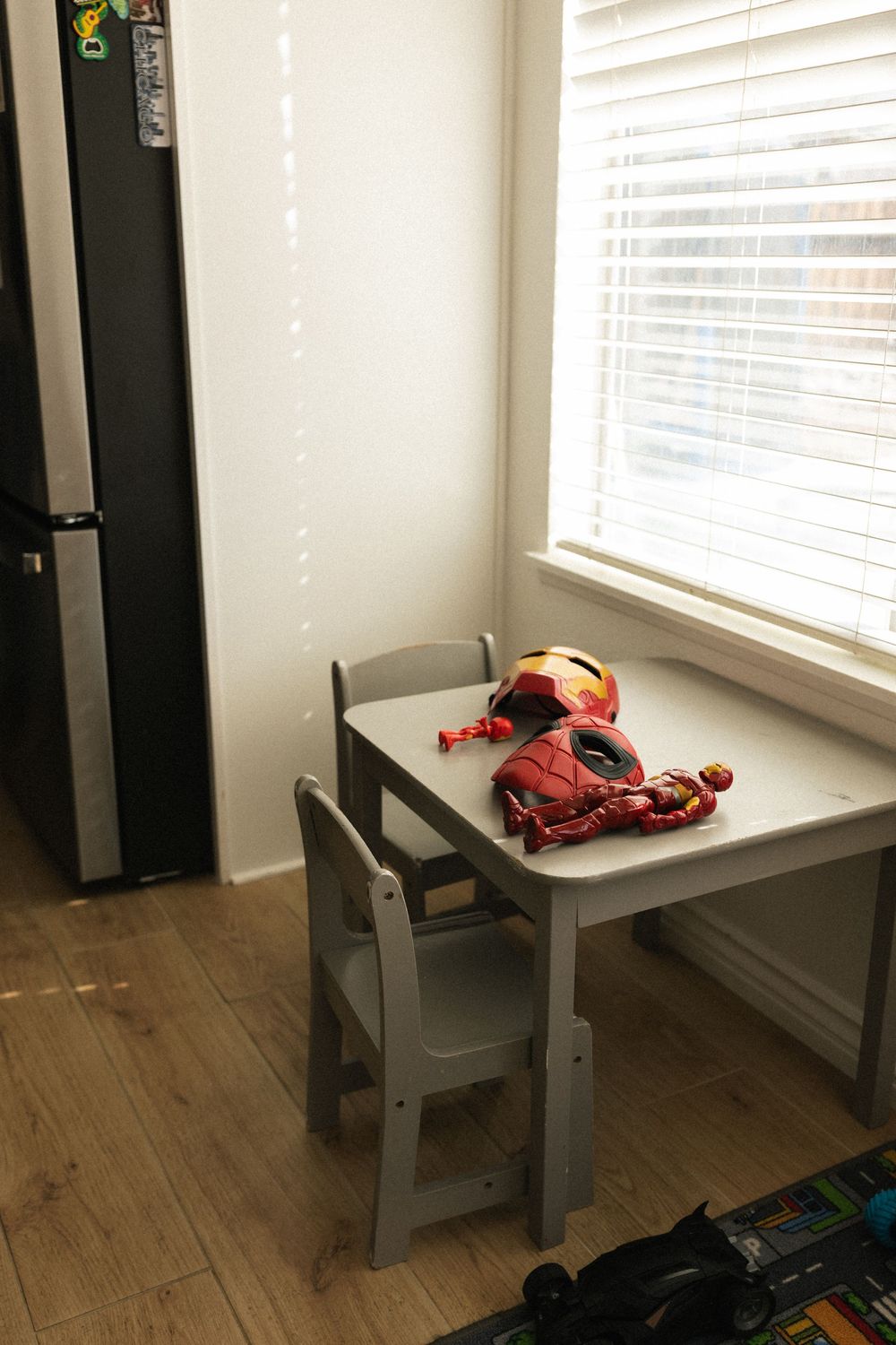 A small gray chair sits empty next to a window with white blinds and wooden flooring.