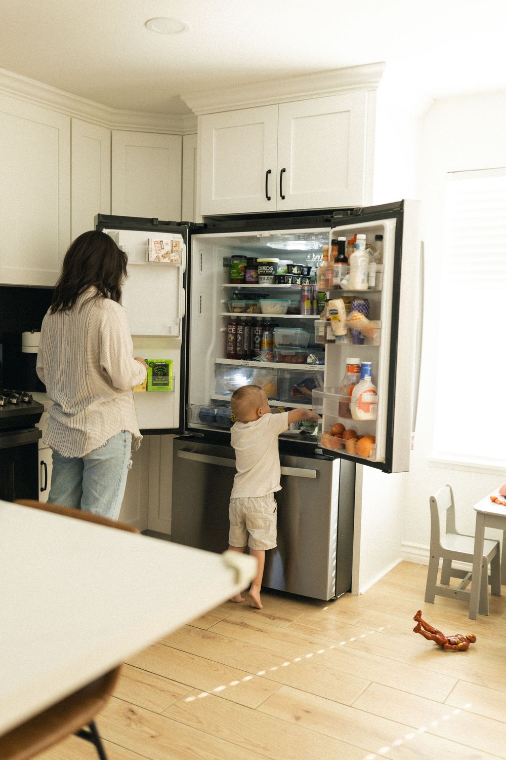 A parent and toddler looking inside an open refrigerator in a bright white kitchen.
