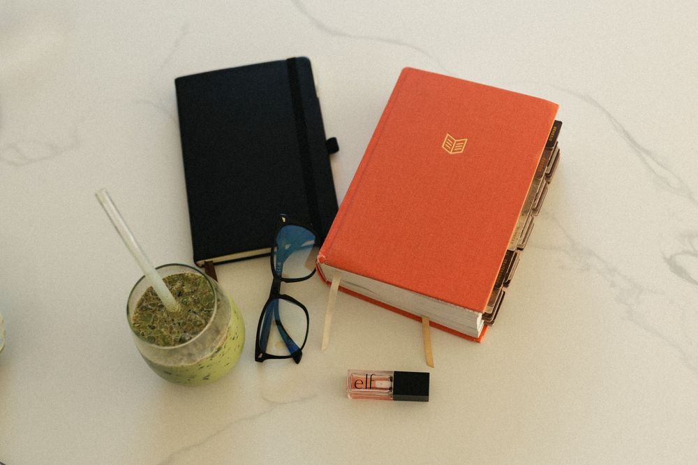 Flatlay of notebooks, reading glasses, lipstick and a green beverage on white marble surface.