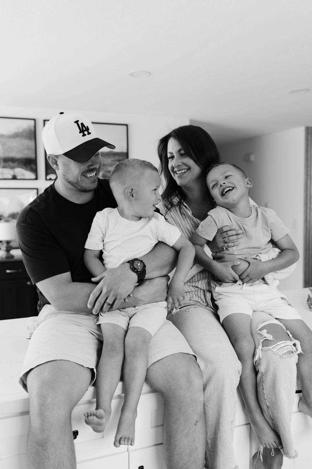A family of four shares joyful laughter while sitting together on a couch in a black and white photograph.
