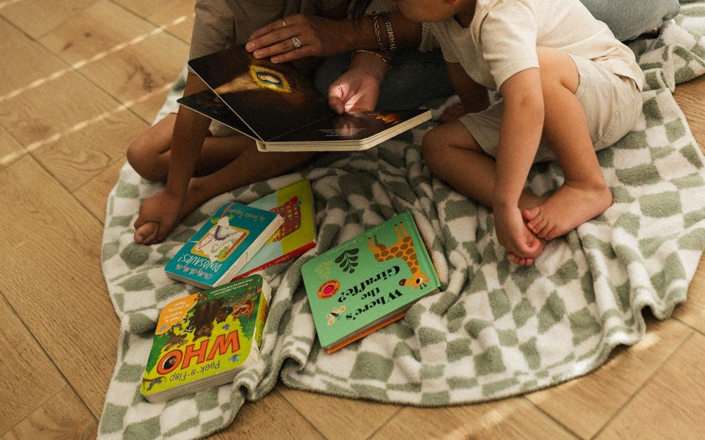 Children reading books while lying on a patterned blanket on tile floor.