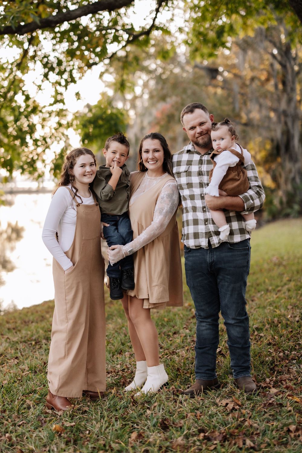 A family group wearing coordinated beige and plaid outfits plays together in fallen autumn leaves during an outdoor photo session.