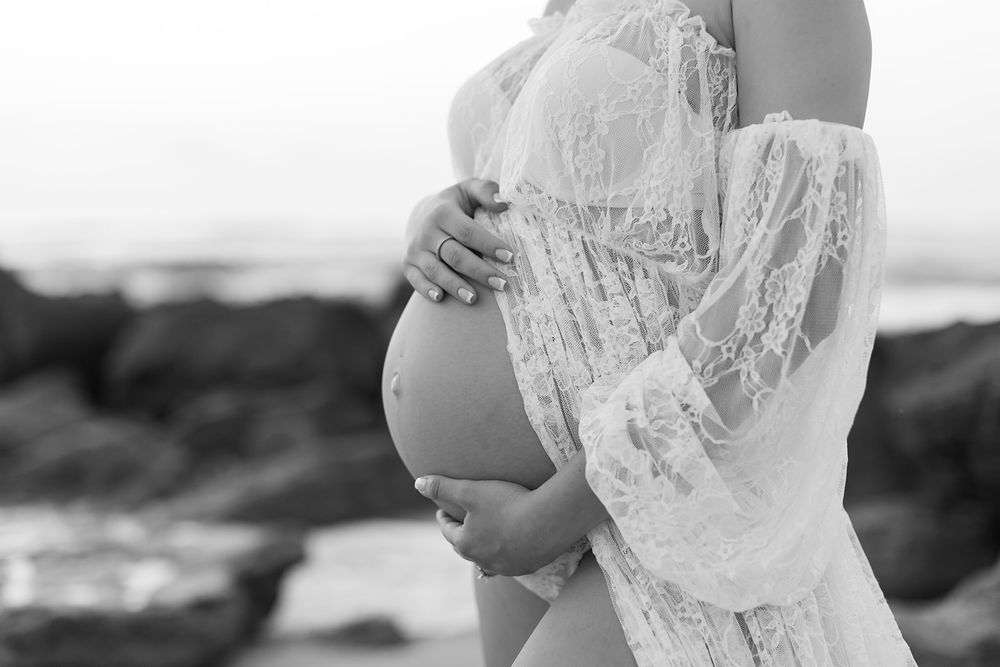 A pregnant woman wearing a flowing white lace dress cradles her belly while standing at a rocky beach in black and white.