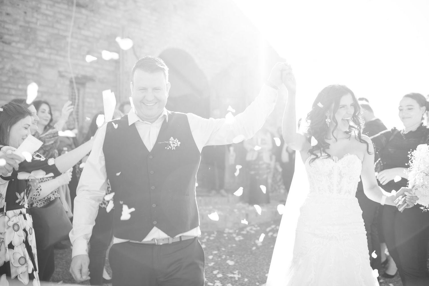 Newlyweds celebrate while guests throw confetti in bright sunlight.