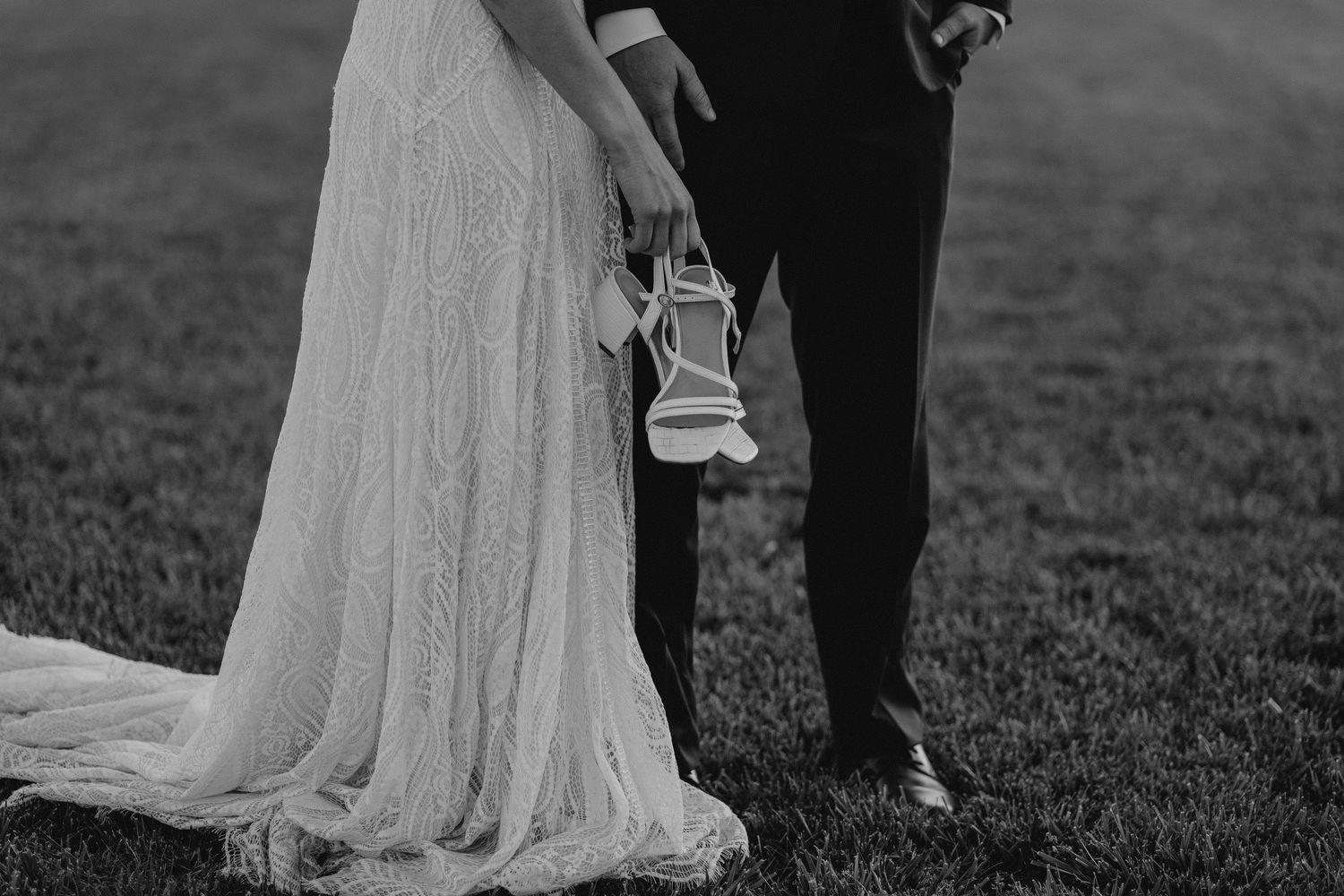 Black and white photo of bride holding her wedding shoes while standing next to groom on grassy field.
