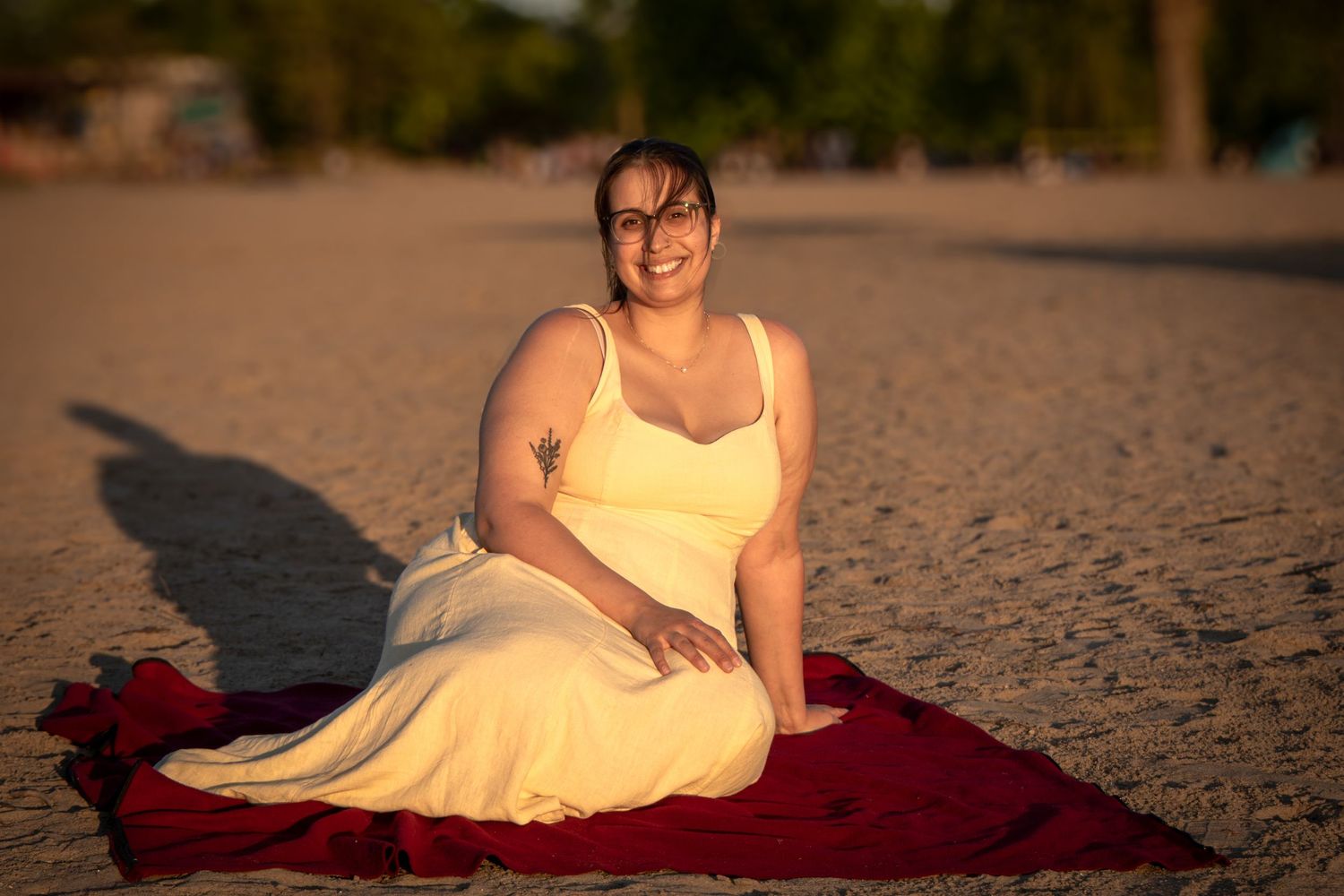 A person in a yellow sundress sits on a red blanket at the beach during golden hour.