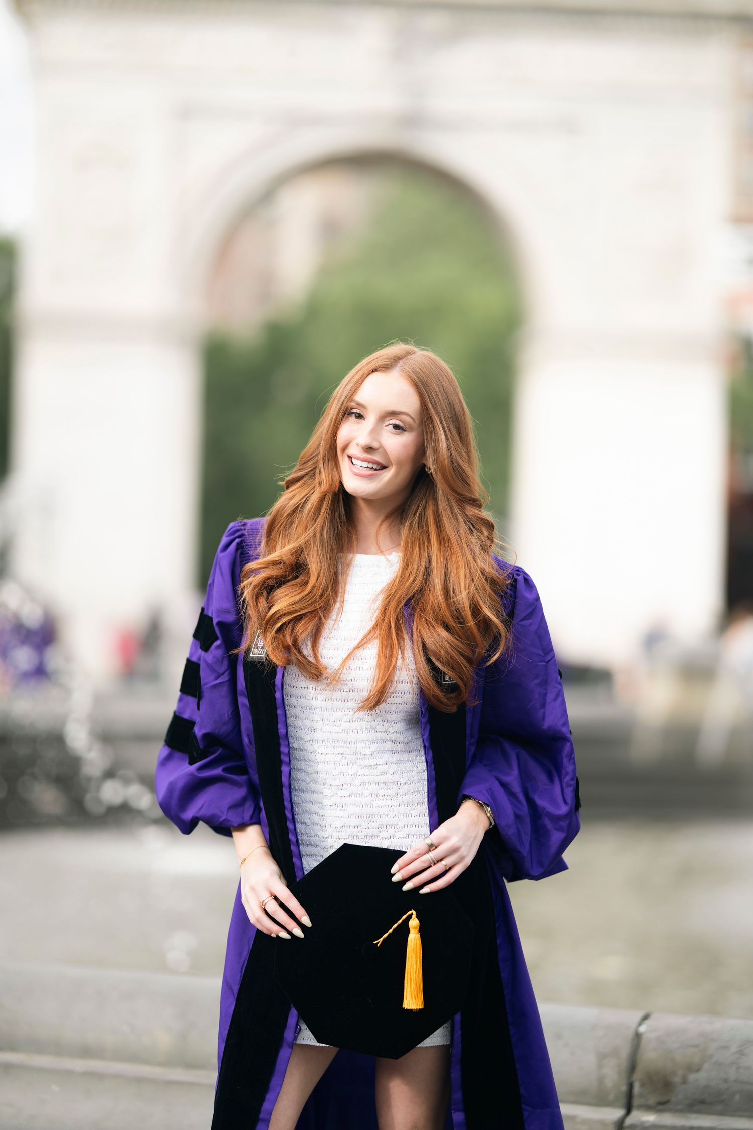 A graduate in a purple graduation robe and cap poses outdoors with a blurred archway in the background.