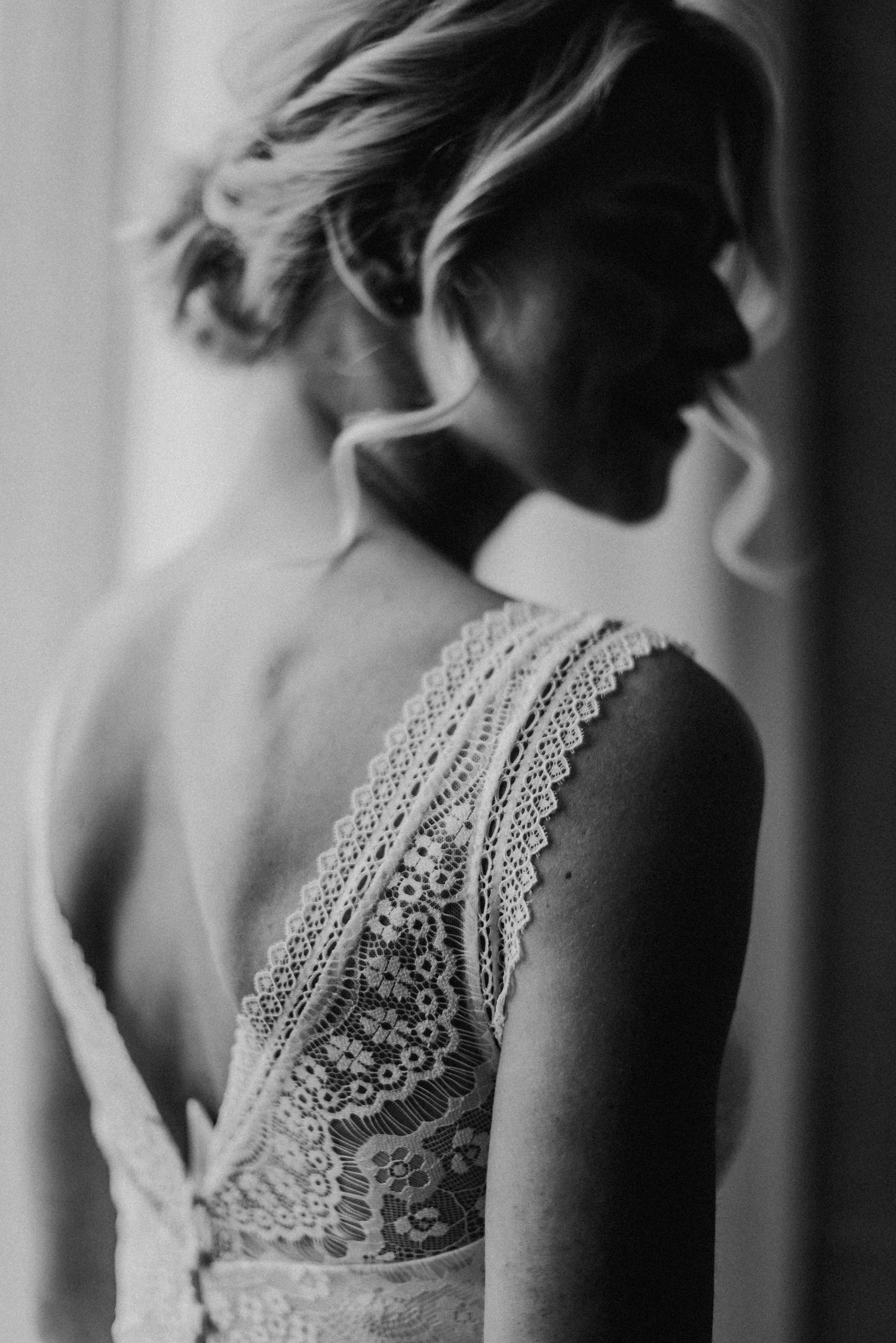 Schwarz-weiß-Foto einer Frau in einem Spitzenkleid, die seitlich steht und aus einem Fenster schaut. Haare sind elegant hochgesteckt.
