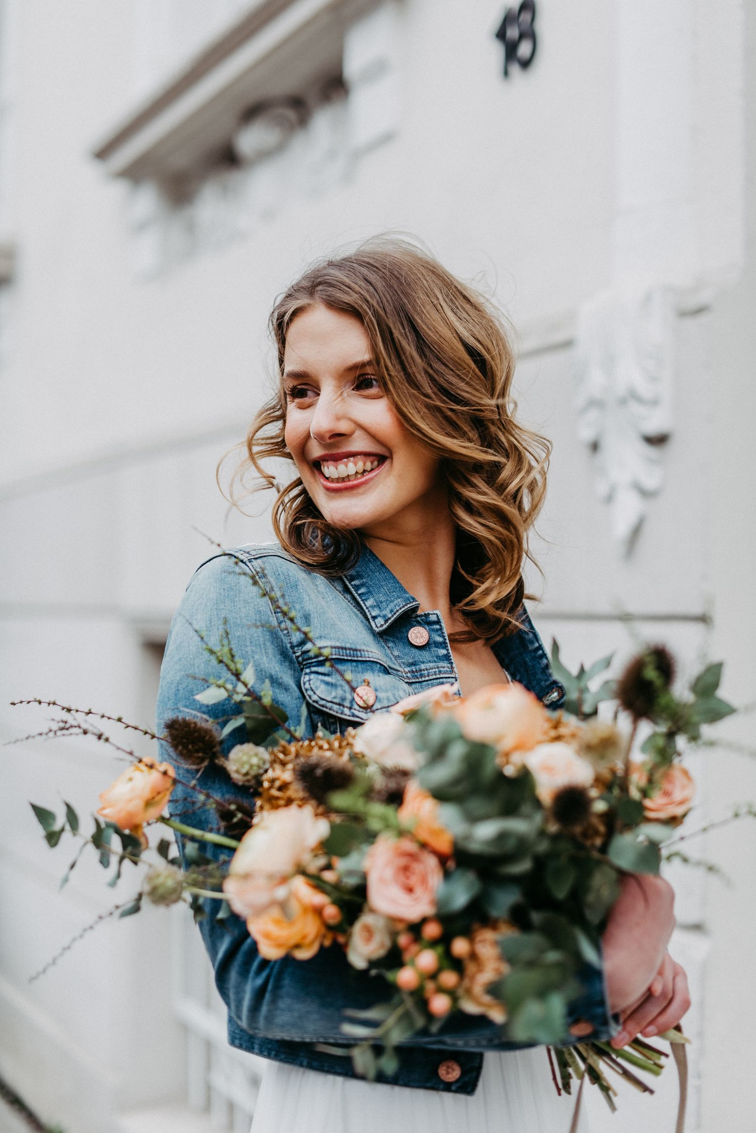 Frau mit lockigem Haar lächelt und hält einen Blumenstrauß aus Rosen und anderen Blumen vor einem Gebäude.