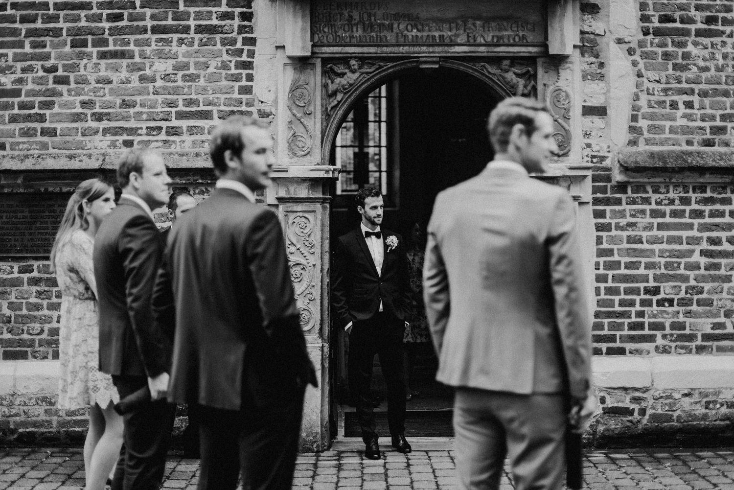 Eine Gruppe von Menschen in formeller Kleidung steht vor einem historischen Gebäude mit Backsteinfassade und verziertem Eingang.
