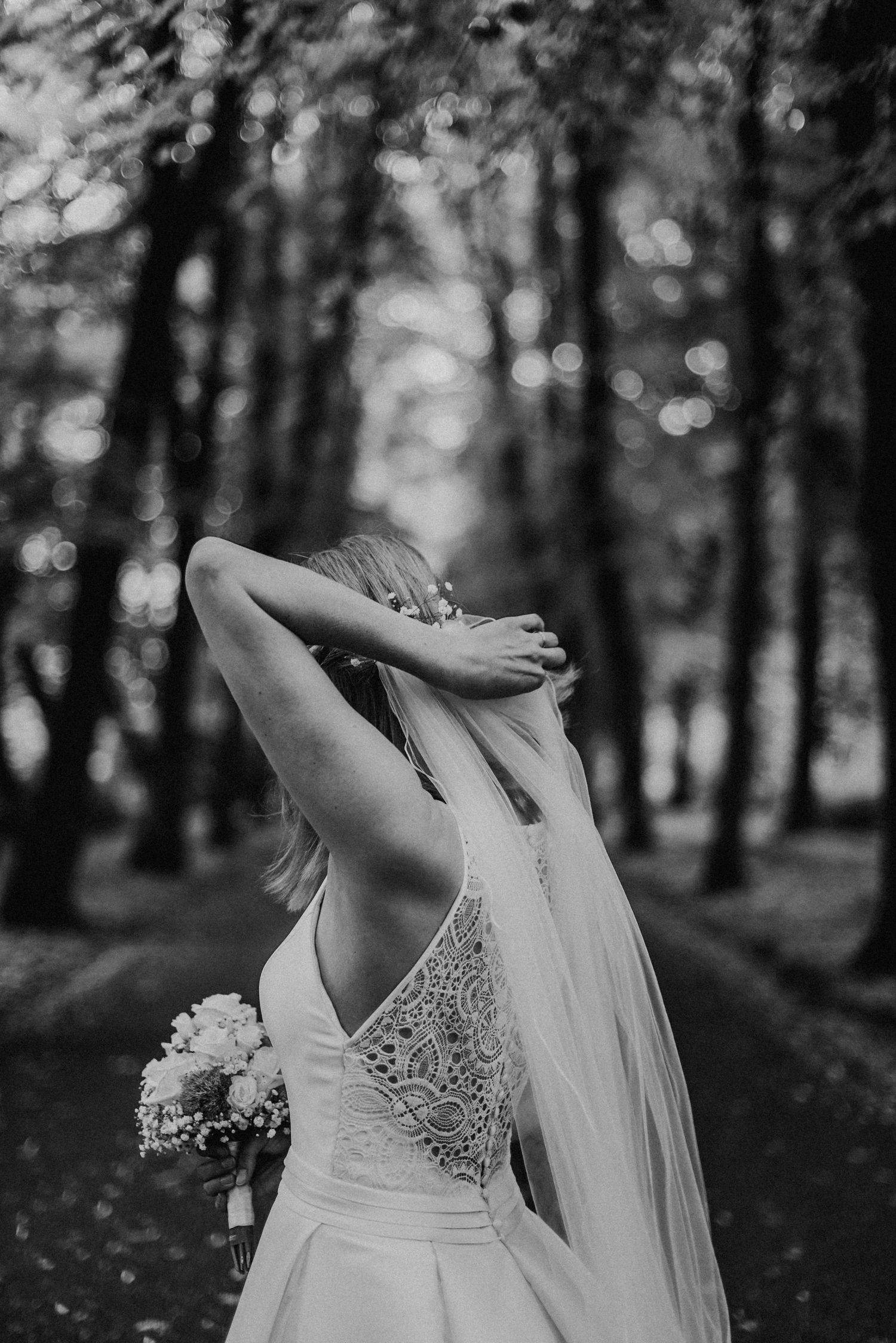 Braut in einem Spitzenkleid steht im Wald, hebt den Arm und hält einen Blumenstrauß. Schwarz-weiß Fotografie.