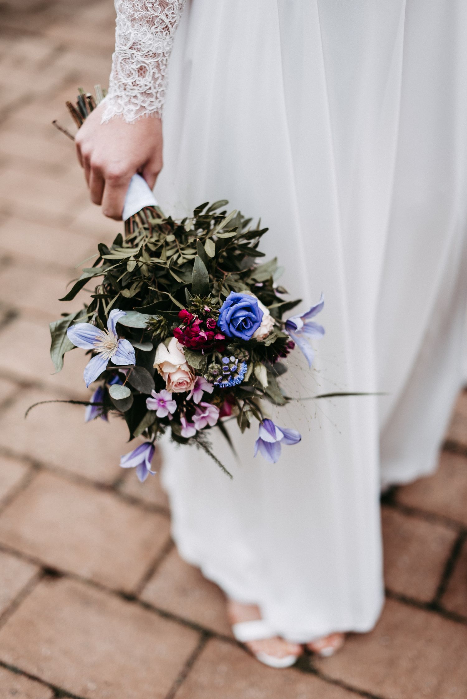 Braut in weißem Kleid hält einen Blumenstrauß mit blauen, lila und roten Blumen auf einem gepflasterten Weg.