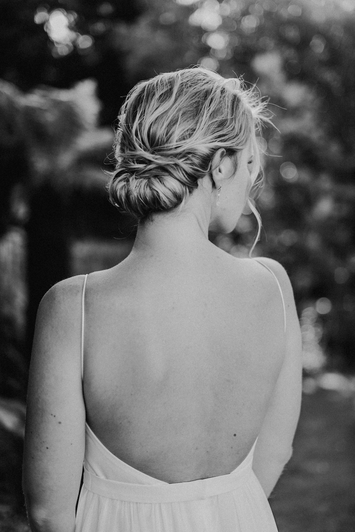 Eine Frau mit blonden Haaren in einem eleganten Dutt trägt ein ärmelloses Kleid mit offenem Rücken, steht im Freien.
