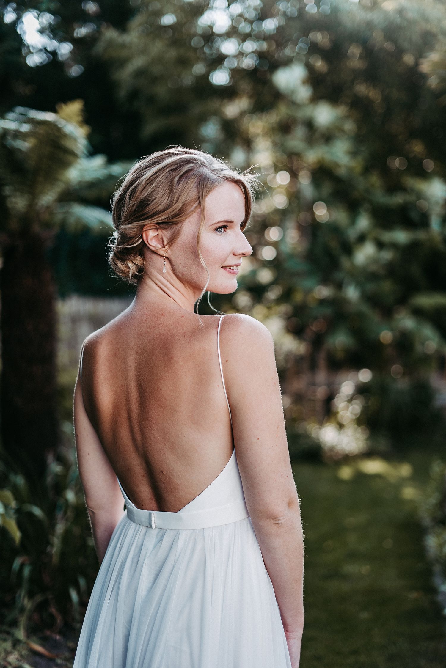 Eine blonde Frau in einem rückenfreihen weissen Kleid steht vor einer Naturkullisse.