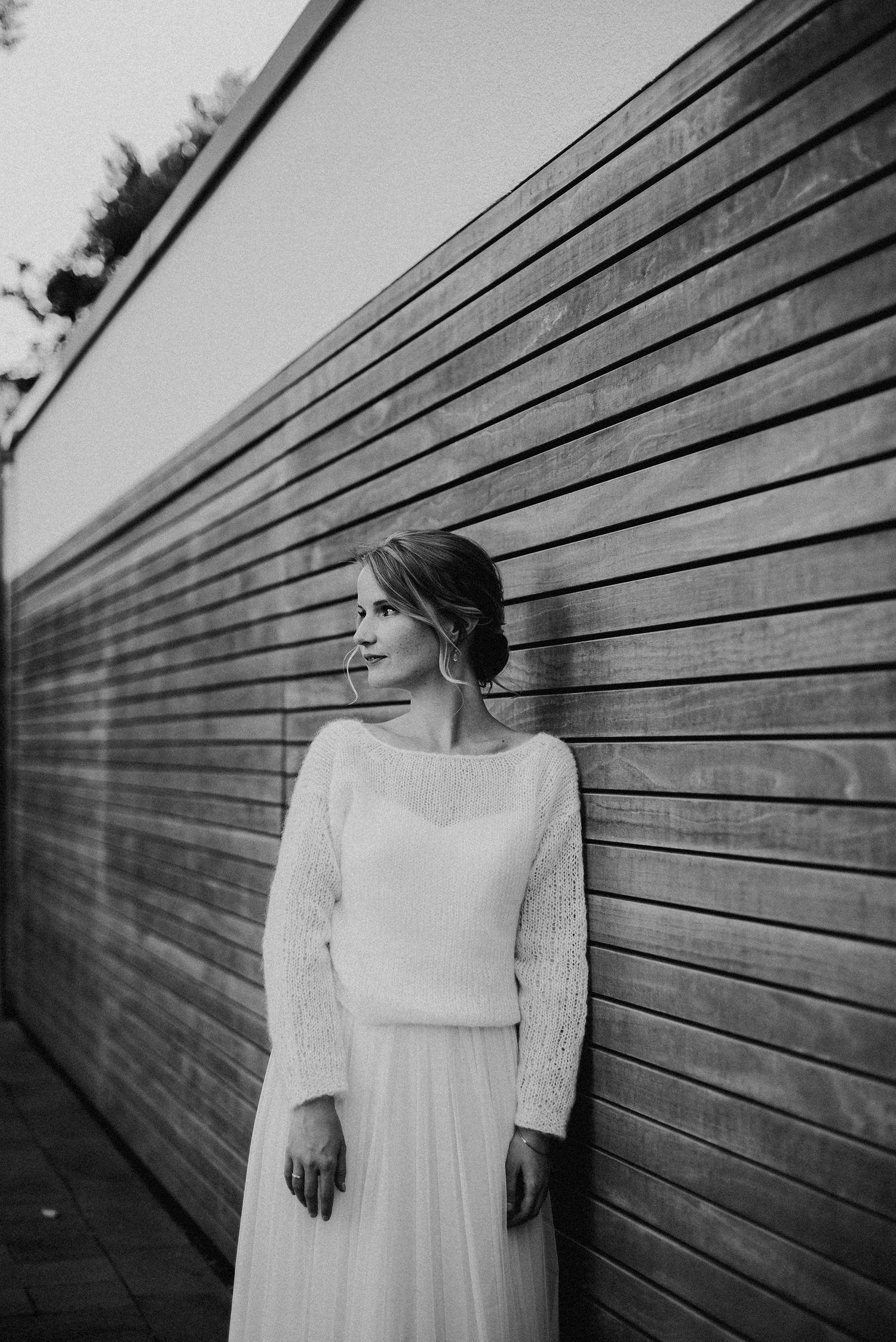 Frau in einem eleganten weißen Kleid lehnt an einer Holzwand. Sie blickt nach links und trägt ihr Haar hochgesteckt.