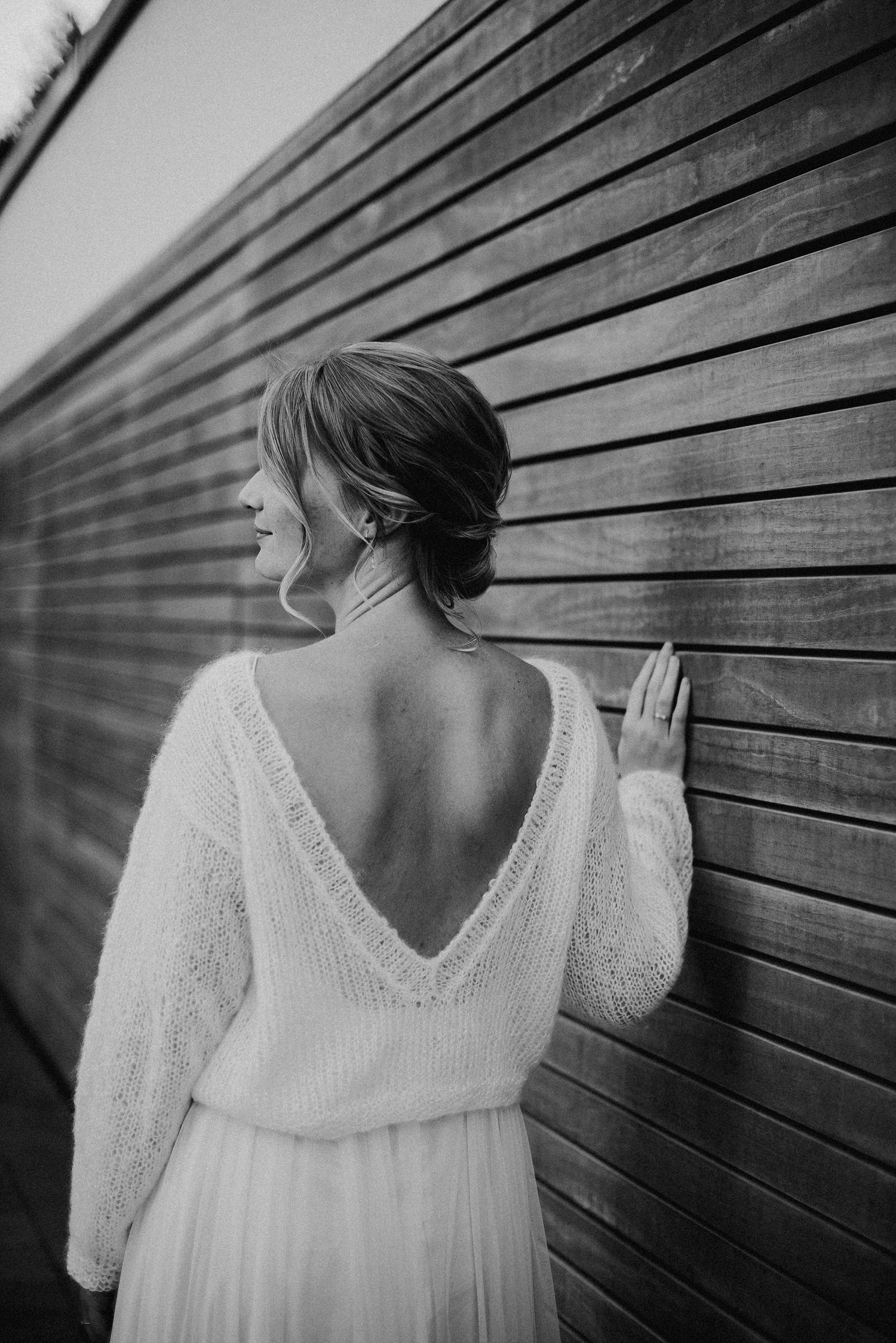 Frau in einem weißen, rückenfreien Pullover lehnt an einer Holzwand. Sie hat hochgestecktes Haar und lächelt leicht.