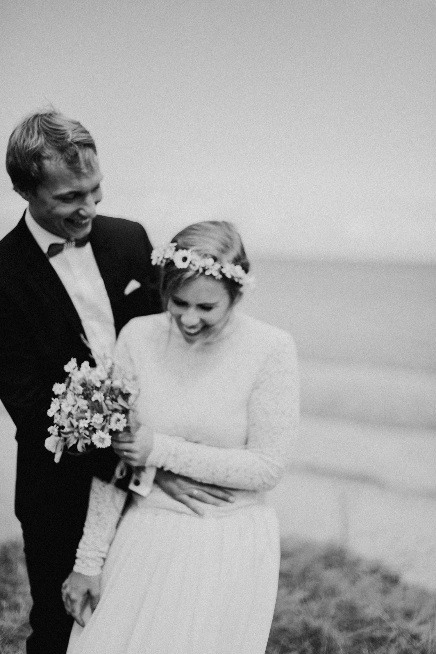 Ein lächelndes Paar in Hochzeitskleidung steht am Strand. Der Bräutigam umarmt die Braut, die einen Blumenstrauß hält.