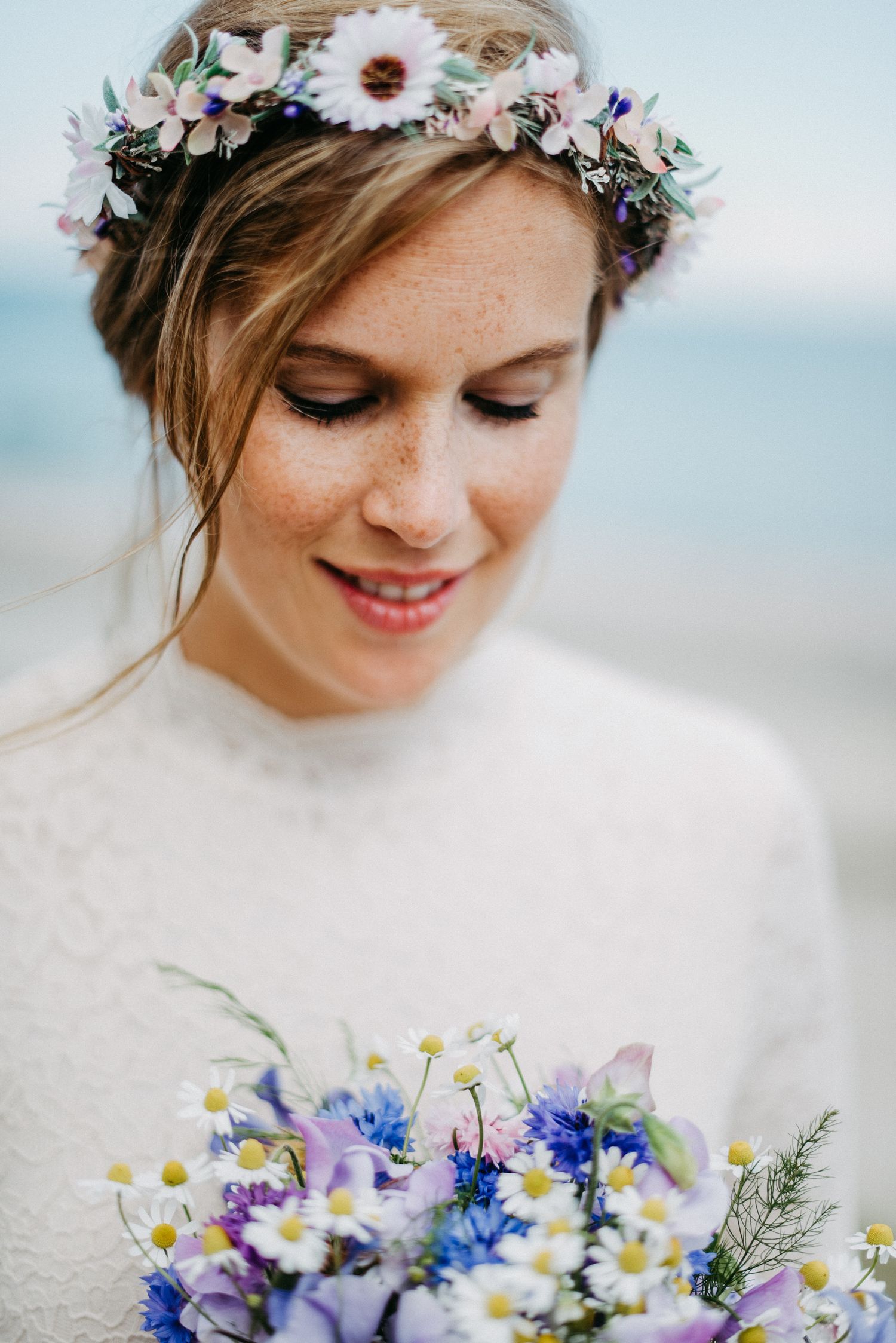 Braut mit Blumenkranz im Haar hält einen bunten Blumenstrauß. Sie trägt ein Spitzenkleid und lächelt sanft.