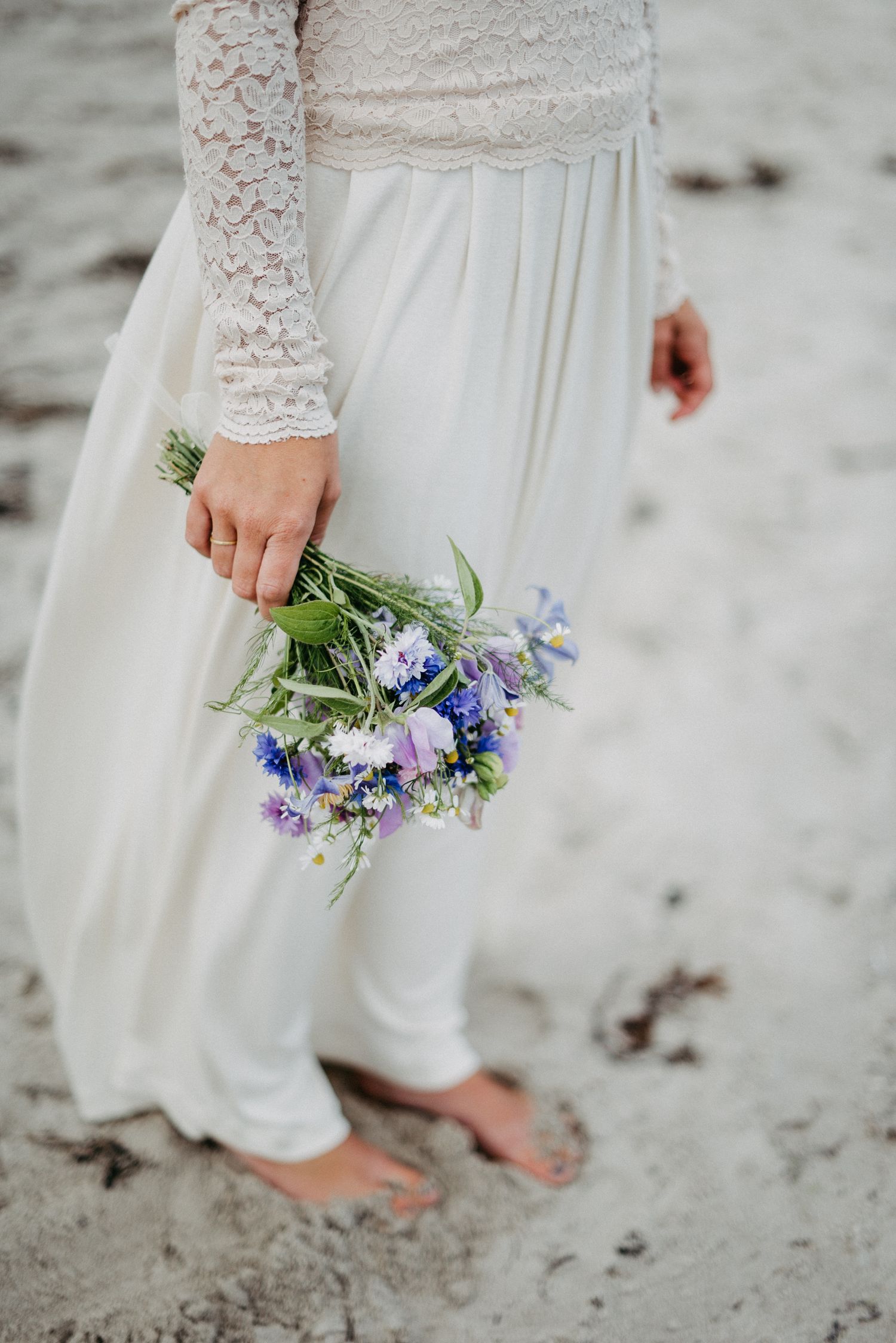 Braut in weißem Spitzenkleid steht barfuß am Strand und hält einen kleinen Blumenstrauß mit lila und weißen Blüten.