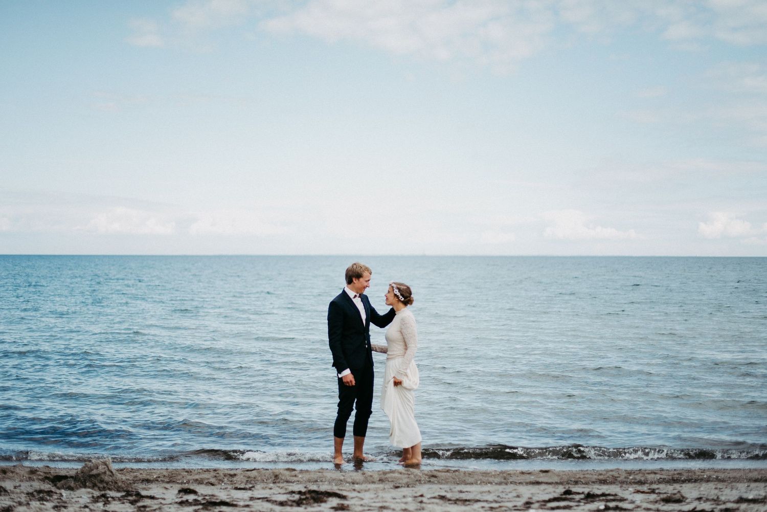 Ein Paar steht am Strand vor dem Meer, beide in eleganter Kleidung. Der Himmel ist leicht bewölkt, und das Wasser ist ruhig.