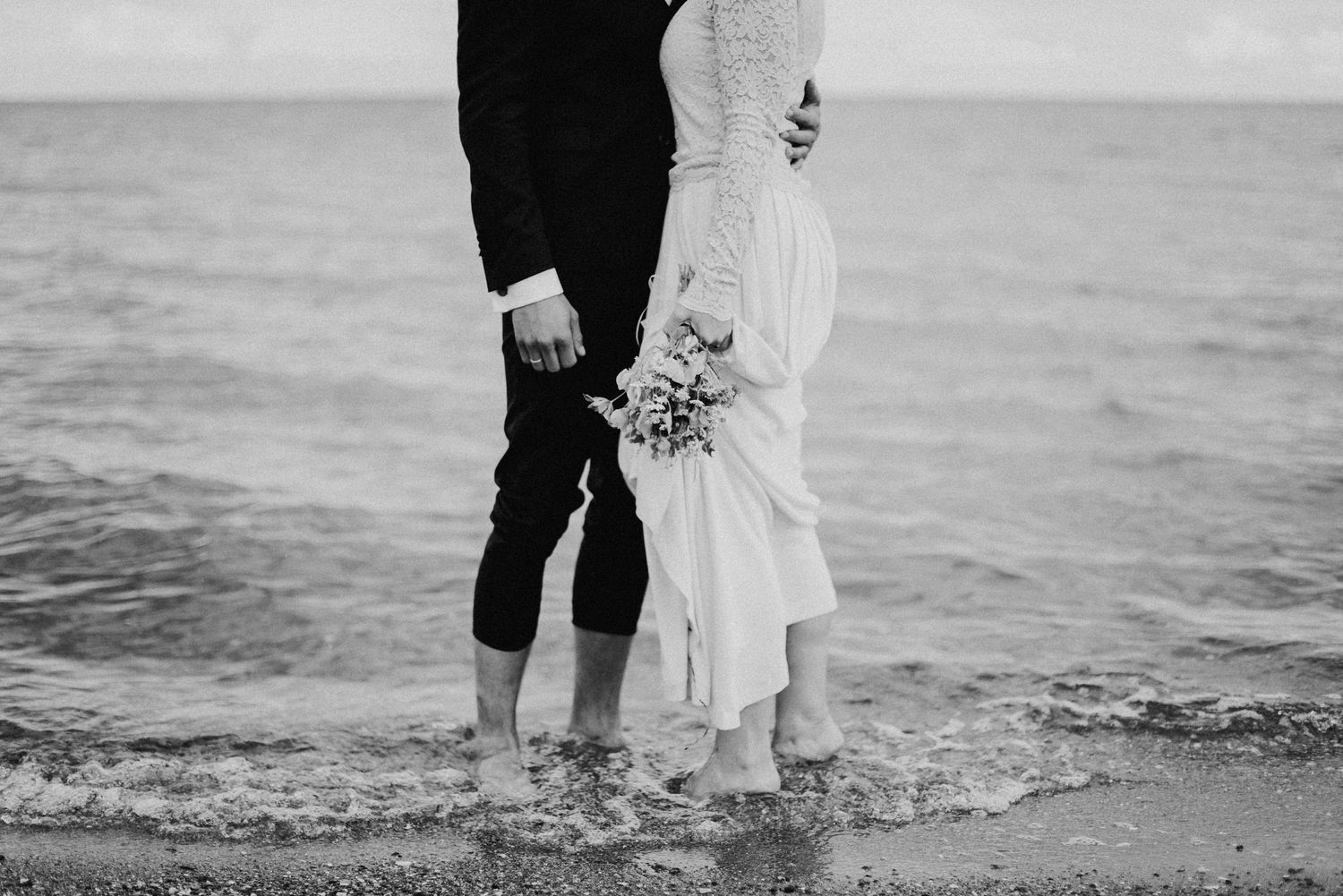 Ein Brautpaar steht mit nackten Füssen im Meer am Strand. Schwarz-Weiss Portrait.