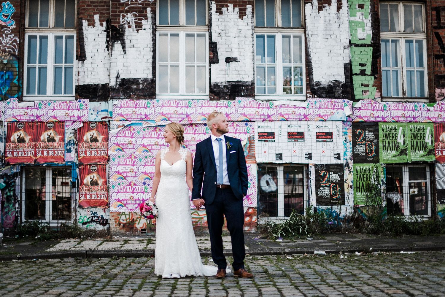Ein Brautpaar steht vor einer bunt bemalten Wand mit Graffiti. Die Braut trägt ein weißes Kleid, der Bräutigam einen Anzug.