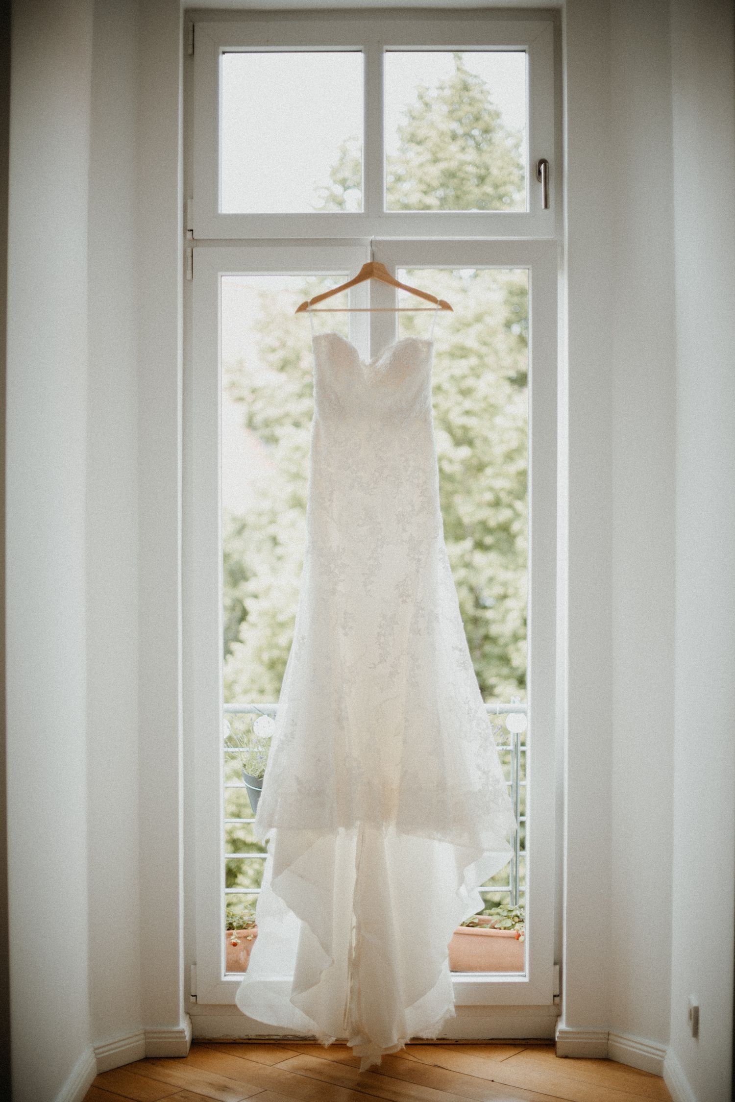 Ein weisses Hochzeitskleid hängt vor einem Fenster, neben dem Fenster hängen weisse Vorhänge. Weiches Licht.
