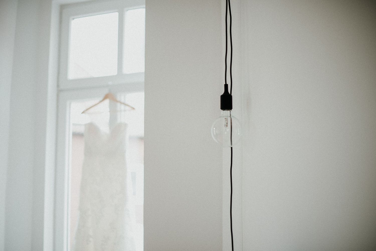 Ein weisses Hochzeitskleid hängt vor einem Fenster, im Vordergrund hängt eine Glühbirne samt Fassung von der Decke. Schwarz-Weiss-Aufnahme.