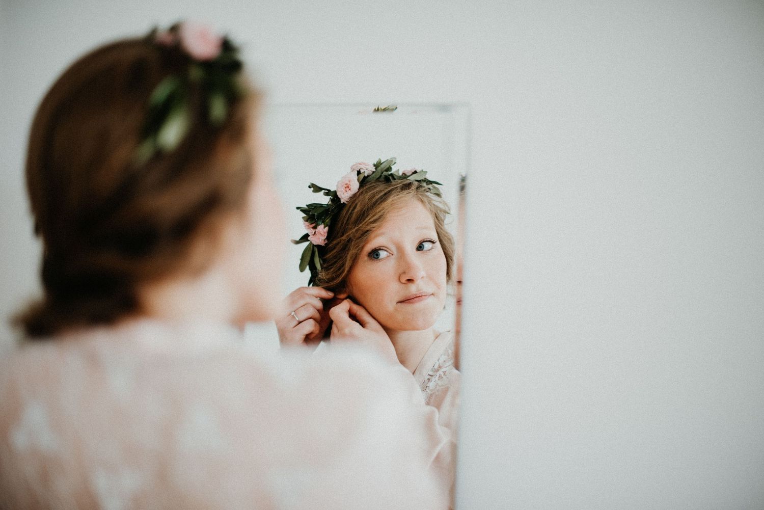 Frau mit Blumenkranz im Haar betrachtet sich im Spiegel und richtet ihre Ohrringe. Sie trägt ein weißes, florales Kleid.