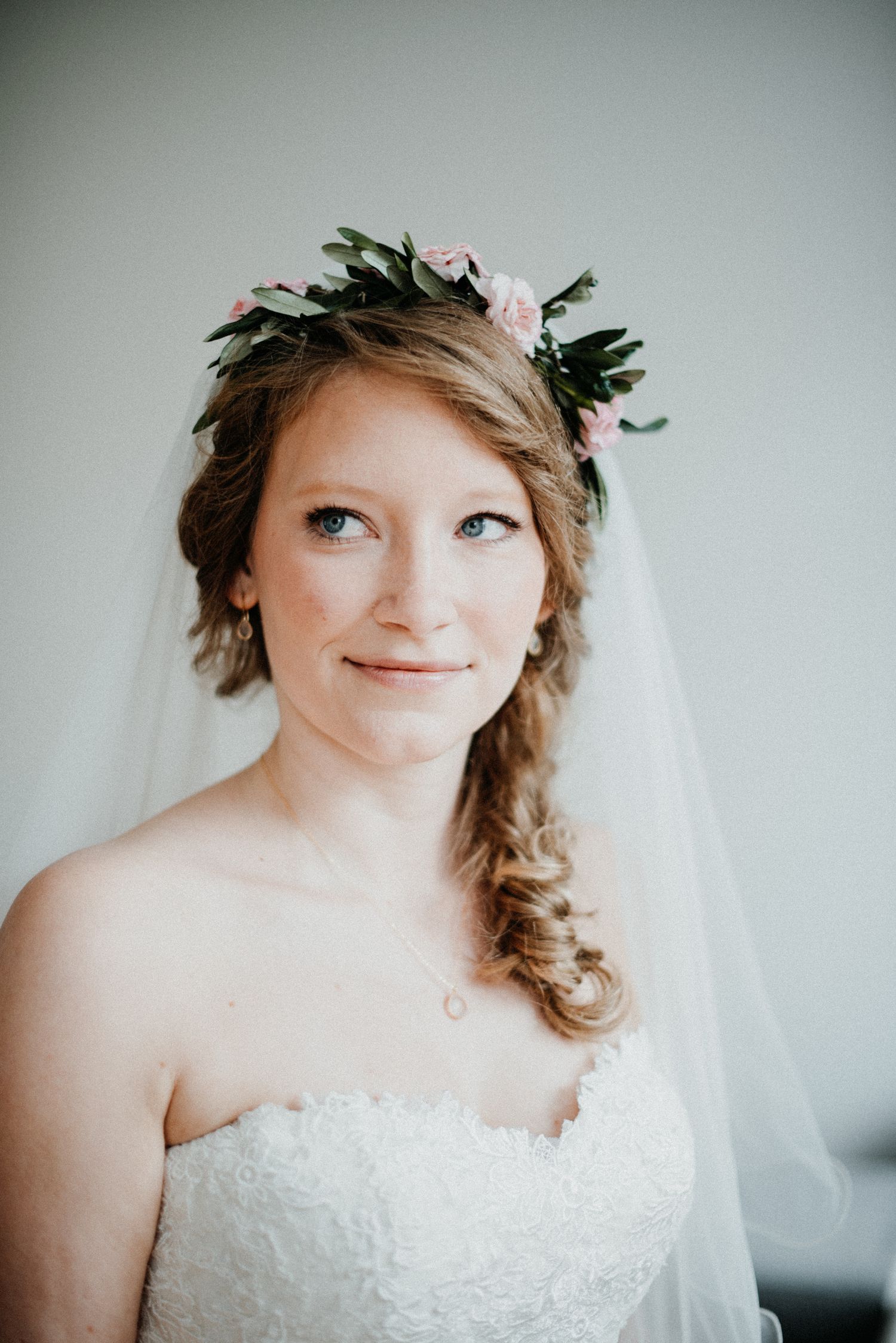 Braut mit Blumenkranz lächelt leicht. Sie trägt ein weißes, schulterfreies Kleid und hat blonde, geflochtene Haare.