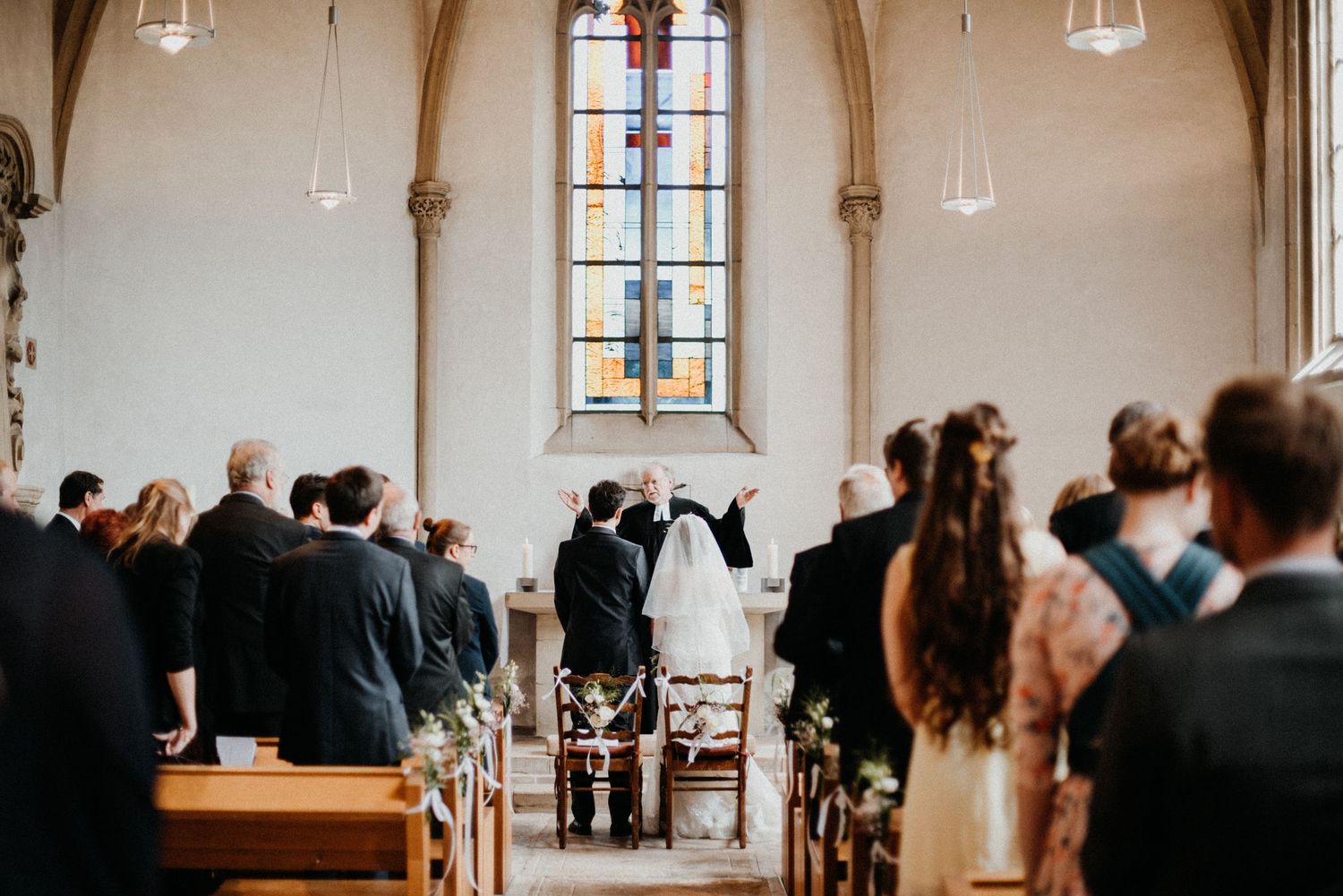 Eine Hochzeitszeremonie in einer Kirche. Gäste sitzen und stehen, während das Brautpaar vor einem bunten Glasfenster steht.