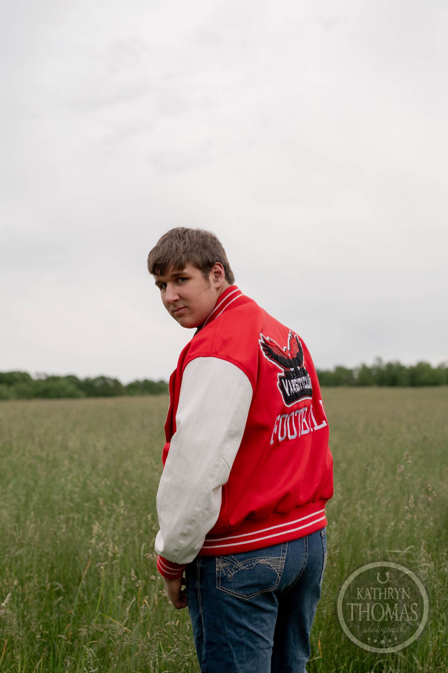 Person wearing a red and white varsity letterman jacket stands in a grassy field on an overcast day.