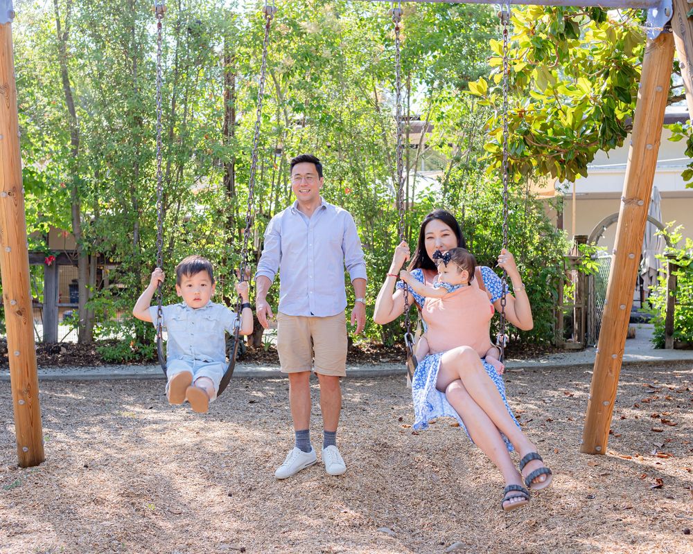 A family enjoys swinging together at a playground on a sunny day in a backyard garden setting.