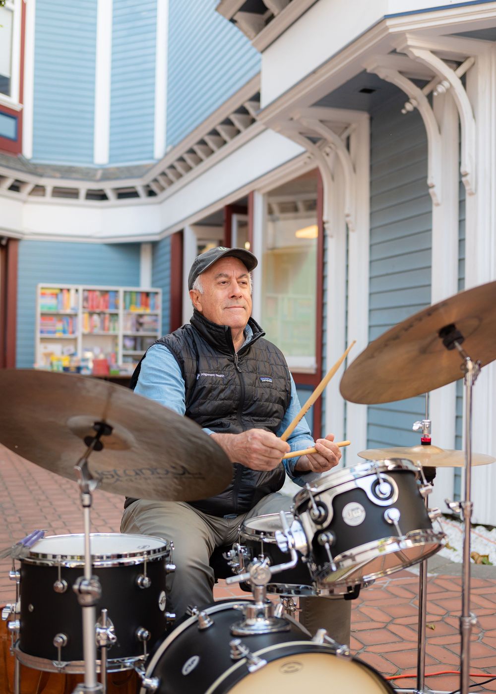Drummer playing on a black drum kit outside a blue-sided building with decorative white trim.