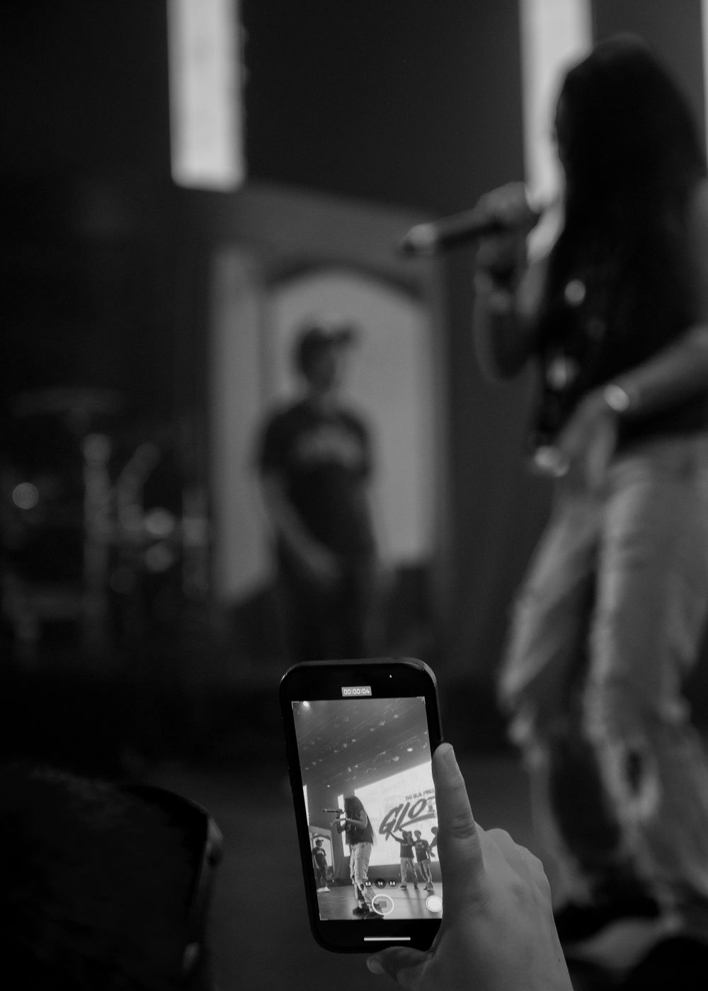 A person holds a phone capturing a concert performance in black and white through the device's screen.