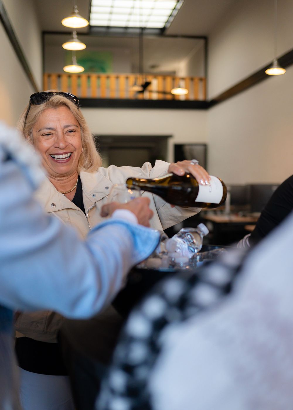 Server pouring wine while smiling warmly in a restaurant dining room with pendant lighting.