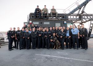 A large group of uniformed personnel and a police dog pose together in front of industrial equipment on a dock.