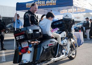 Police motorcycle unit parked at outdoor event with onlookers gathered around in sunny daytime setting.