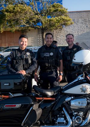 Three police officers stand smiling next to their motorcycles and patrol car under a tree.