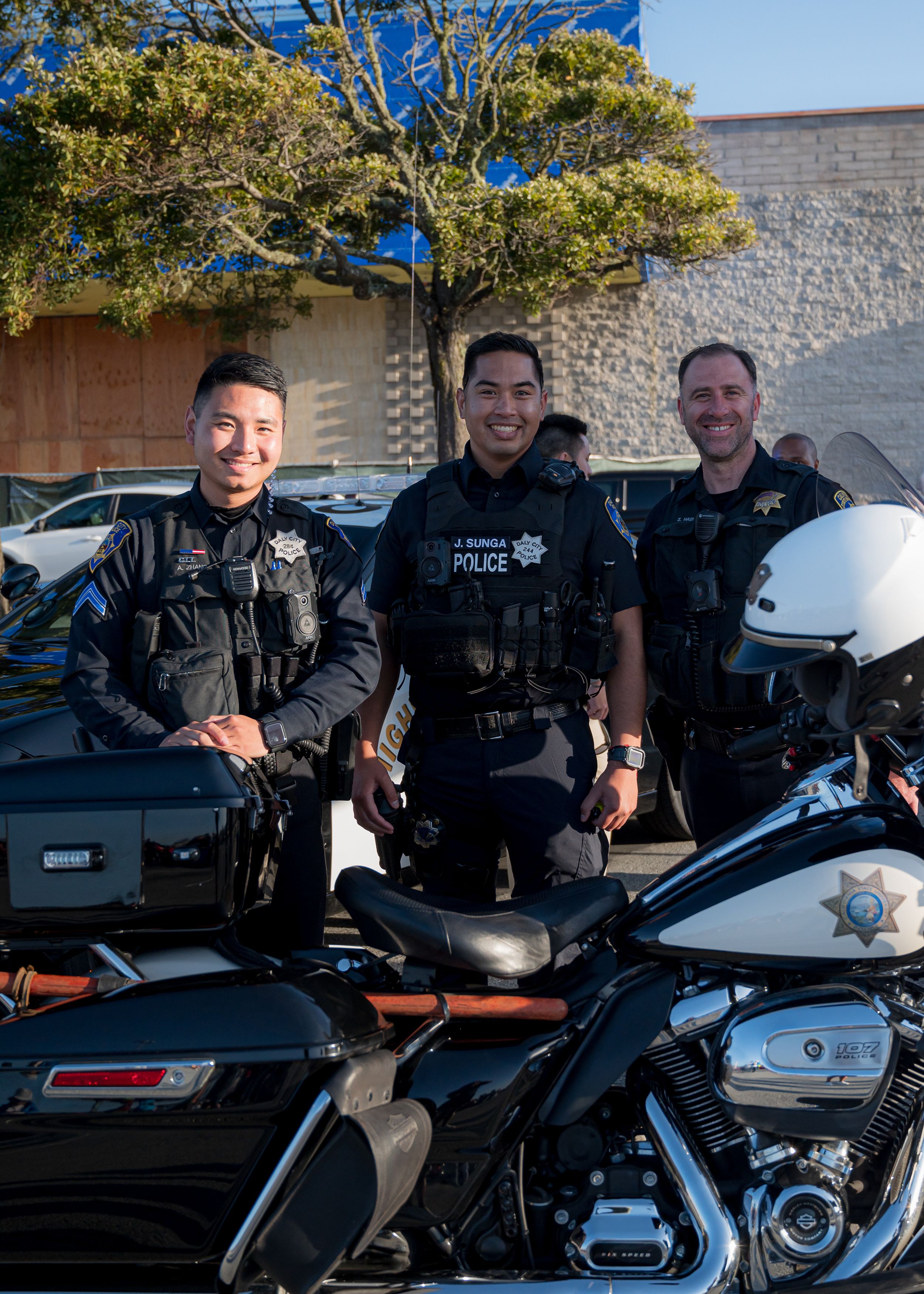 Three police officers stand smiling next to their motorcycles and patrol car under a tree.