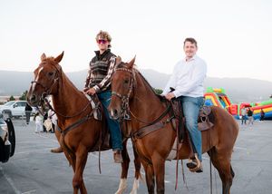 Two riders sit atop brown horses at an outdoor event with mountains in the background.