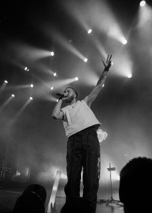 Dramatic black and white concert photo of performer raising peace sign on stage with spotlights beaming through smoke.